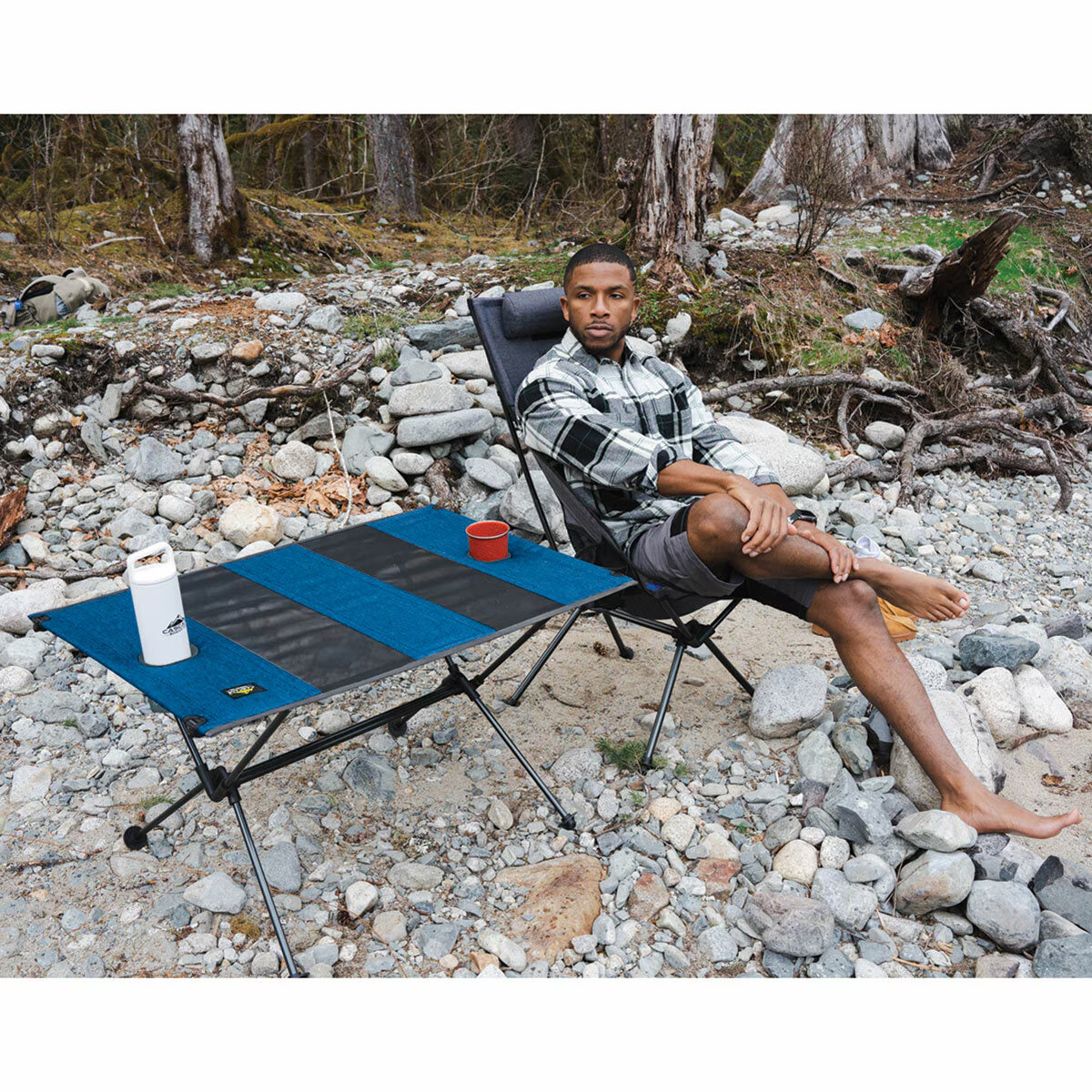 Man relaxing in portable camping chair with foldable table on rocky riverbank, ideal for van life and outdoor adventures