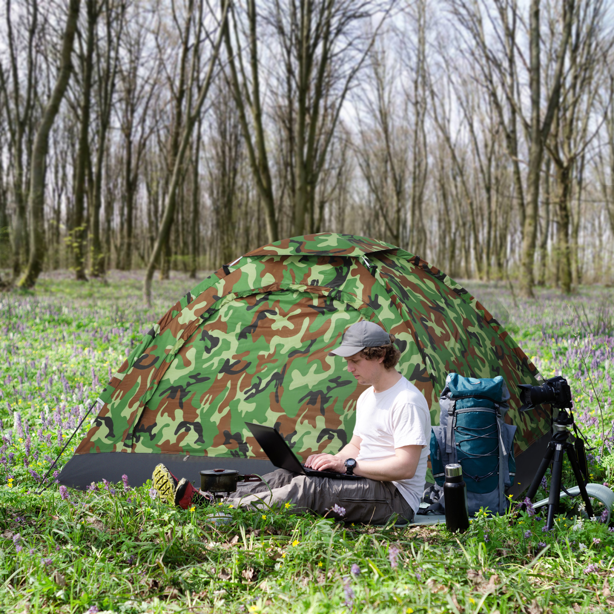 Man using laptop by camo tent in wildflower field, camping gear nearby for van life adventures