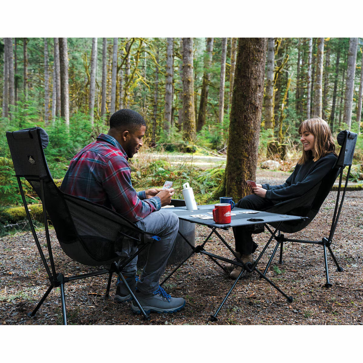 Couple playing cards at portable camping table with foldable chairs in forest, ideal for van life adventures