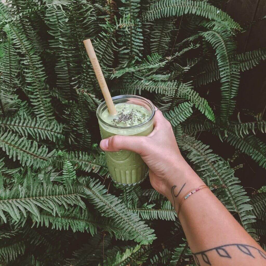 Hand holding green smoothie in glass jar with straw, surrounded by lush ferns, perfect for van life adventures