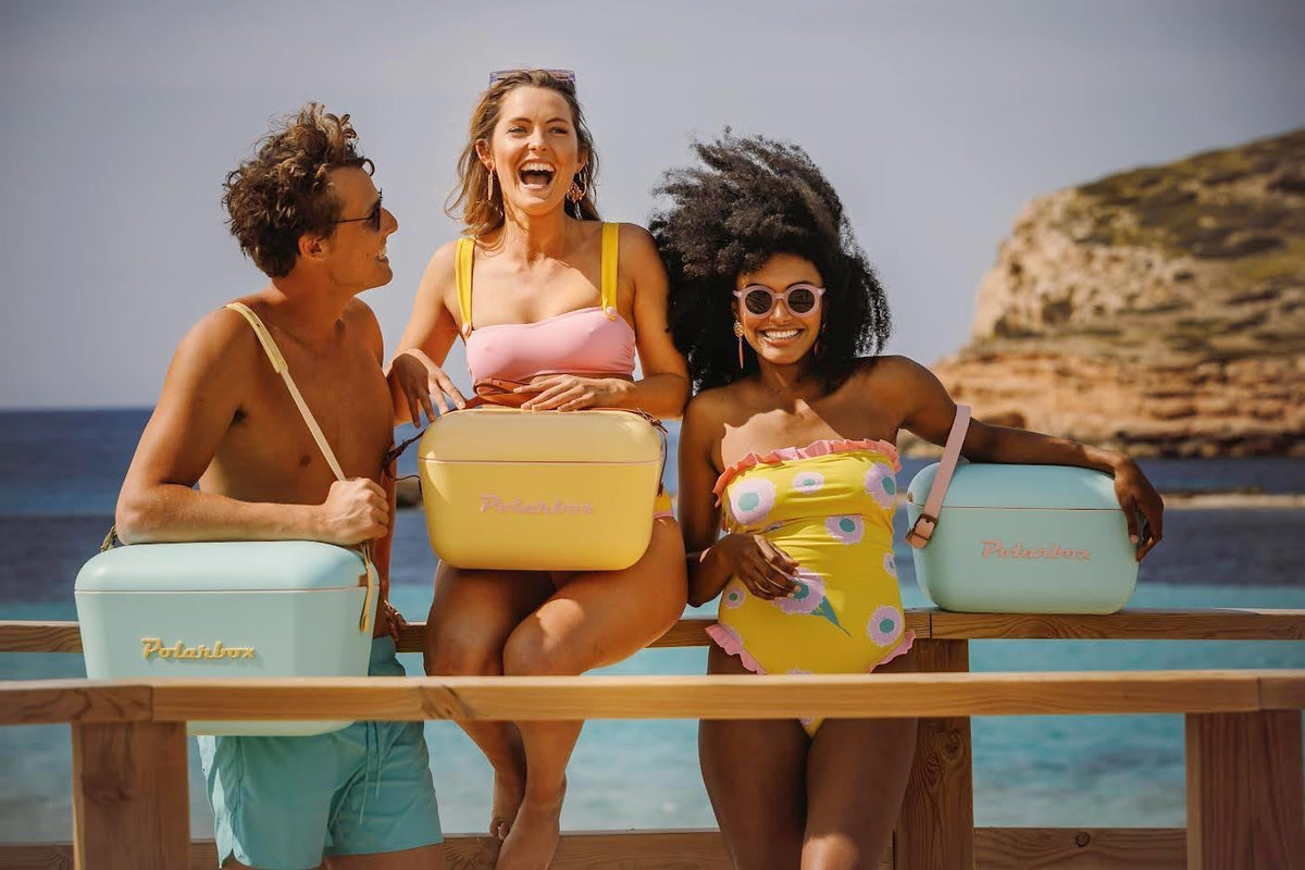 Three friends in swimwear laugh on a wooden railing by the sea, each holding a pastel-coloured insulated box with shoulder straps.