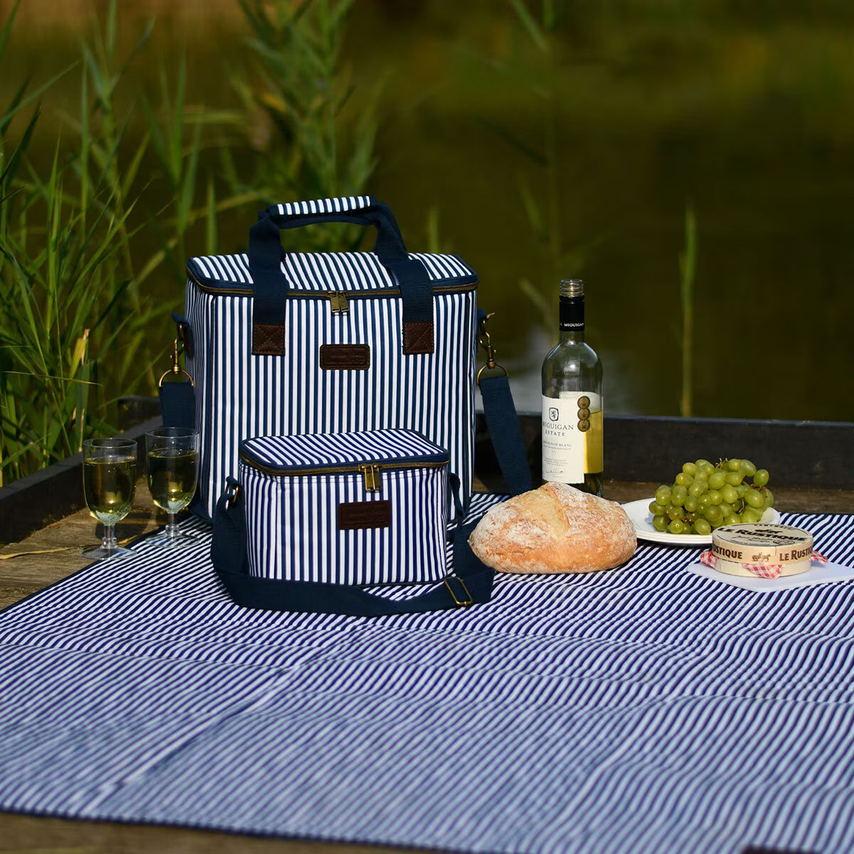 Navy and white striped picnic cooler bags with matching blanket, wine, bread, and cheese by a lakeside