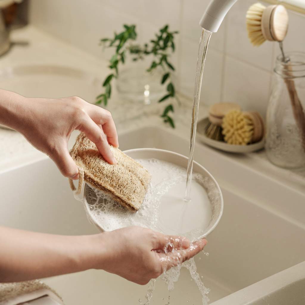 Hand washing dish with natural loofah sponge under running water in kitchen sink, eco-friendly van life essential
