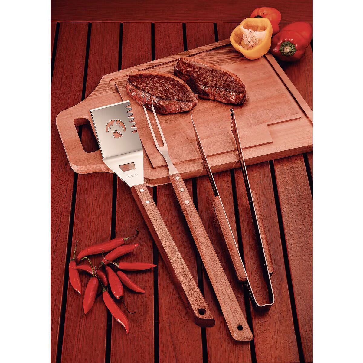 Stainless steel BBQ tools with wooden handles, steaks, peppers on cutting board for outdoor grilling