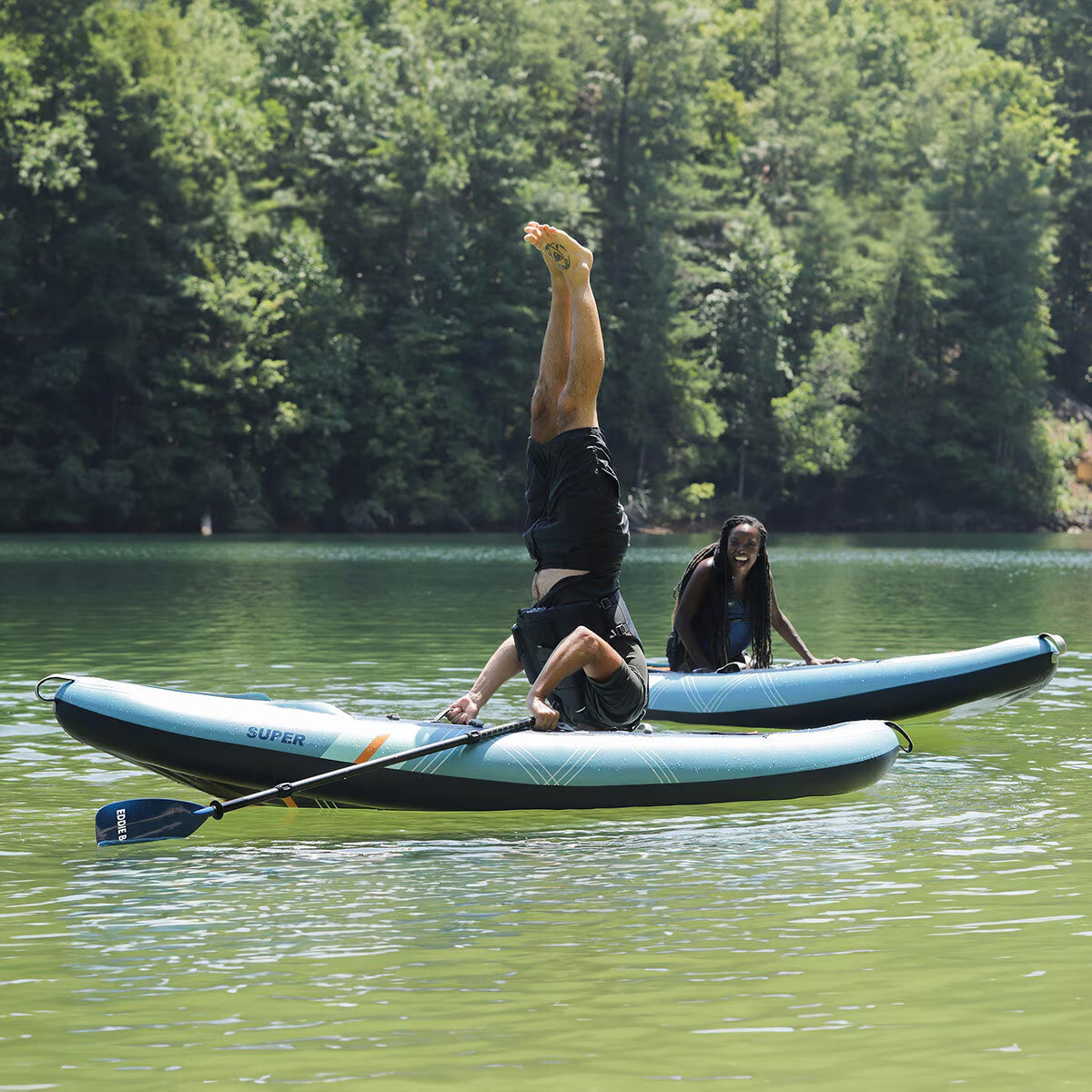 Man doing handstand on inflatable SUP paddleboard with woman laughing nearby on lake, outdoor adventure gear