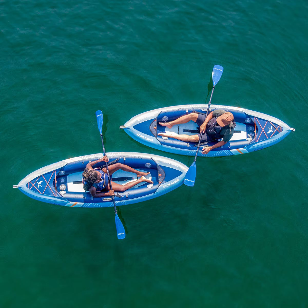 Two people kayaking in blue Eddie Bauer inflatable kayaks on calm turquoise water, perfect for outdoor adventures.